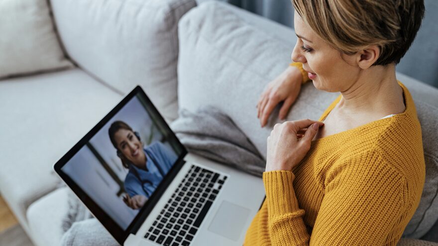 A woman on her couch on a telehealth video visit with a doctor on her laptop